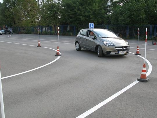 Szary samochód nauki jazdy marki Opel z eMką na dachu podczas manewru jazdy po łuku na placu manewrowym wyznaczonym pachołkami i tyczkami