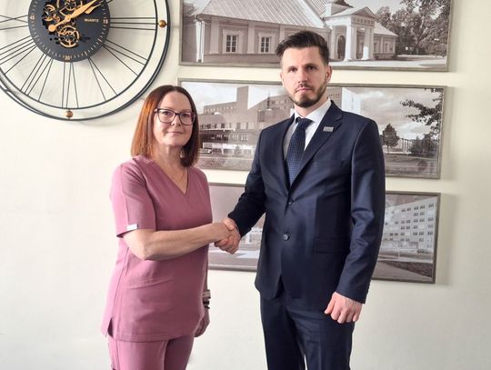 Mężczyzna w ciemnym garniturze i kobieta w różowym stroju medycznym podają sobie ręce w geście porozumienia. Stoją w jasnym pomieszczeniu na tle ściany, na której wisi duży ozdobny zegar z widocznym mechanizmem oraz czarno-białe zdjęcia architektury szpitalnej w ramkach.