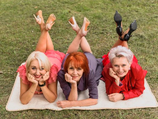 Trzy uśmiechnięte kobiety w różnym wieku, ubrane w eleganckie, kolorowe stroje (różowy, fioletowy, czerwony), leżą wspólnie na kocu na trawie, patrząc z radością w stronę obiektywu.