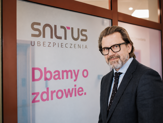 Robert Łoś, Prezes Zarządu SALTUS Ubezpieczenia, stojący w okularach i garniturze na tle szklanych drzwi z logo SALTUS Ubezpieczenia i hasłem Dbamy o zdrowie