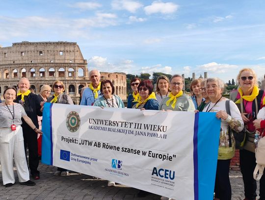Grupa seniorów stoi na rzymskim placu, trzymając duży biały baner z logo Akademii Bialskiej i napisem „UTW AB Równe szanse w Europie”. W tle widać monumentalną, antyczną architekturę Koloseum pod błękitnym niebem. Uczestnicy mają na szyjach charakterystyczne żółte chusty.