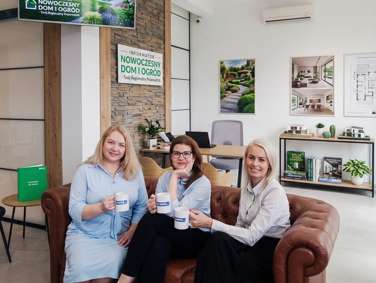 Trzy uśmiechnięte kobiety siedzą na brązowej, pikowanej kanapie w nowoczesnym biurze. Trzymają białe kubki z logo. W tle widać jasne wnętrze, biurko oraz elementy promocyjne informatora „Nowoczesny Dom i Ogród”. Atmosfera profesjonalizmu i otwartości.