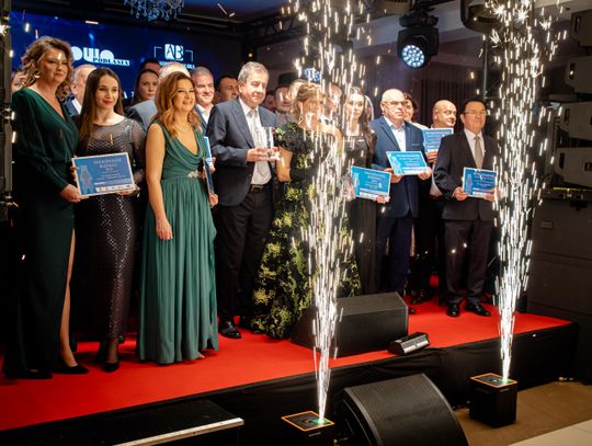 Grupa elegancko ubranych kobiet i mężczyzn stoi na scenie z czerwonym dywanem, trzymając w rękach dyplomy i statuetki. W tle widać logo Słowa Podlasia, a na pierwszym planie z podłogi strzelają dwa efektowne, iskrowe fontanny (zimne ognie).
