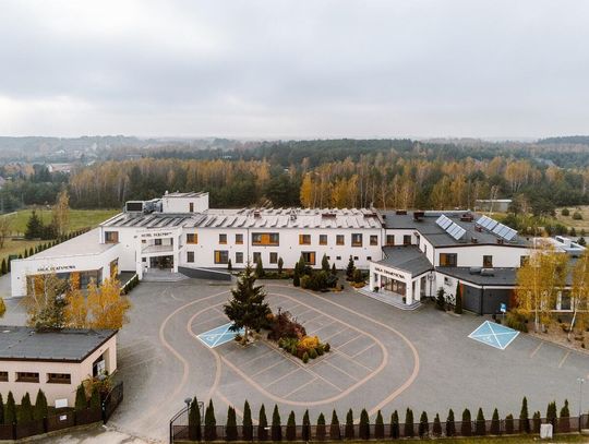 Zdjęcie z lotu ptaka przedstawiające nowoczesny budynek Hotelu Polonia*** w Grabanowie, z widocznym głównym wjazdem, parkingiem oraz otaczającym go zalesionym terenem.