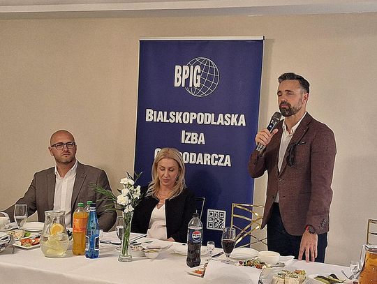 Trzy osoby przy stole prezydialnym podczas oficjalnego spotkania. Po prawej stronie stoi mężczyzna w brązowej marynarce z mikrofonem w ręku i przemawia. Na środku siedzi blondynka w czarnym żakiecie, a po lewej stronie siedzi mężczyzna w okularach i szarej marynarce.