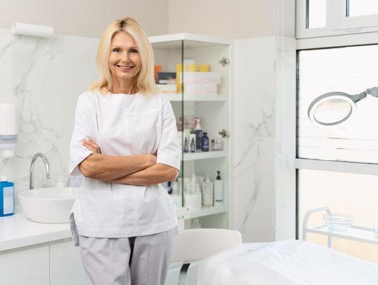 Uśmiechnięta kosmetolog w białym fartuchu stoi w jasnym gabinecie. W tle przeszklona witryna z kosmetykami, zlew oraz profesjonalna lampa zabiegowa. Zdjęcie buduje wizerunek profesjonalizmu i higieny w branży beauty, promując cykl „Wiosenne Odświeżenie”.