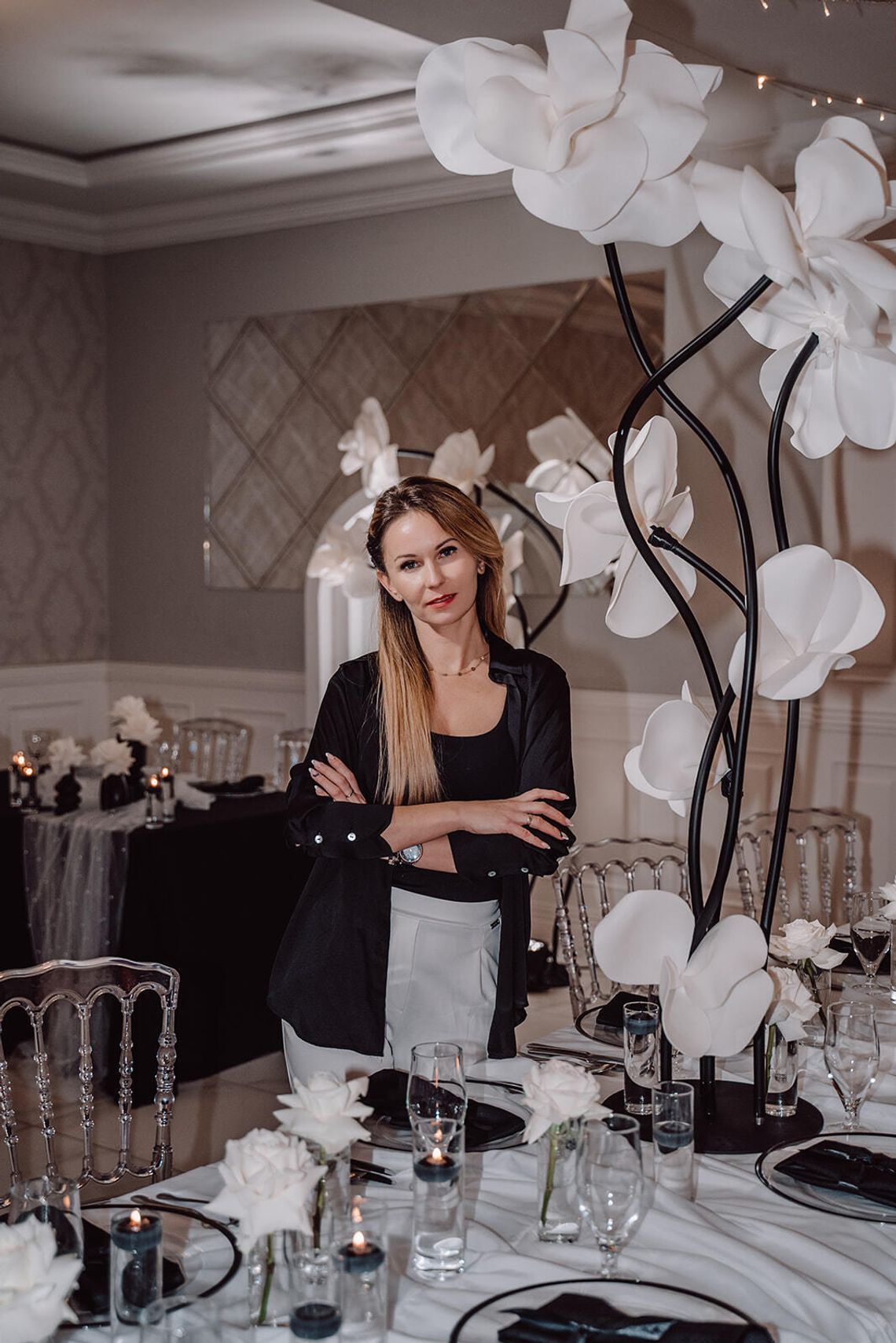 CARMEN FLOR flowers&deco Portret kobiety stojącej w elegancko przystrojonej sali bankietowej, otoczonej kompozycjami kwiatowymi, w tym dużymi dekoracyjnymi białymi kwiatami na stojakach i stołem z białymi różami.