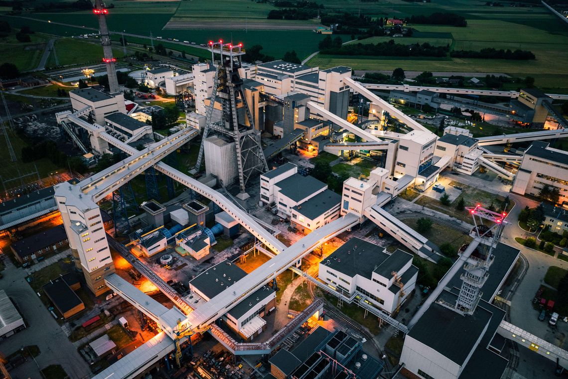 Widok z lotu ptaka na przemysłowy kompleks kopalni węgla kamiennego LW Bogdanka, z widocznymi wieżami szybowymi i systemem taśmociągów, oświetlony w godzinach wieczornych.