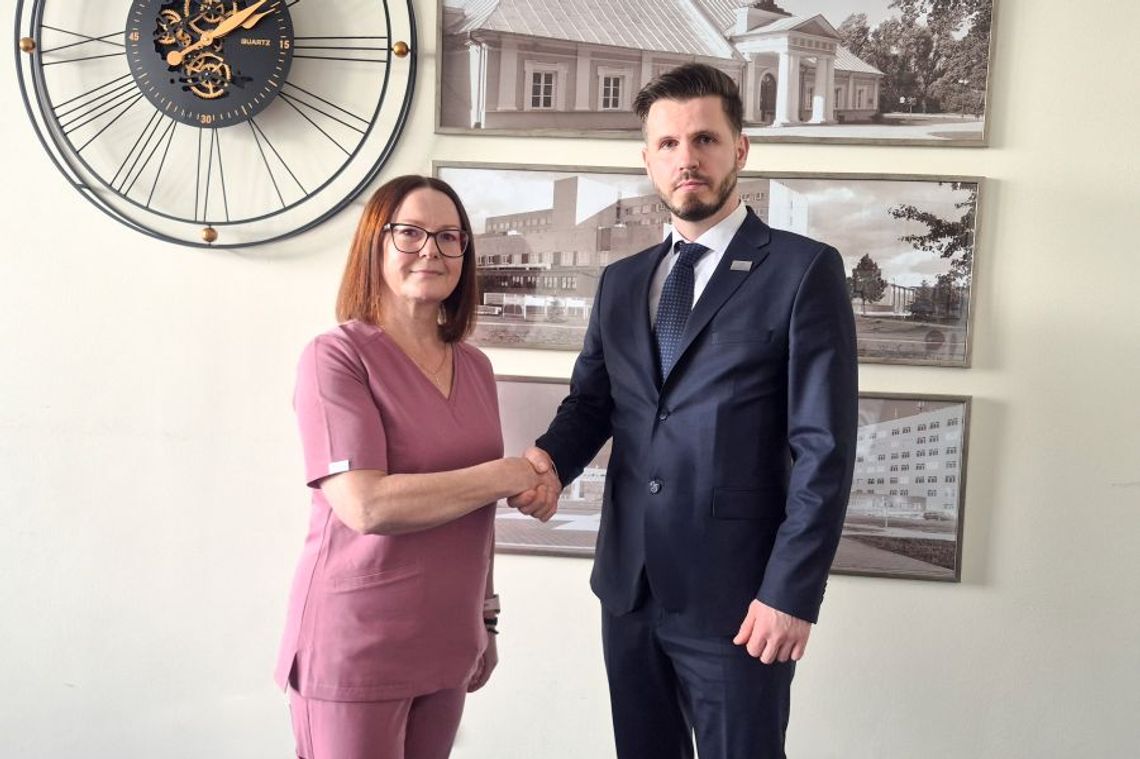 Mężczyzna w ciemnym garniturze i kobieta w różowym stroju medycznym podają sobie ręce w geście porozumienia. Stoją w jasnym pomieszczeniu na tle ściany, na której wisi duży ozdobny zegar z widocznym mechanizmem oraz czarno-białe zdjęcia architektury szpitalnej w ramkach.