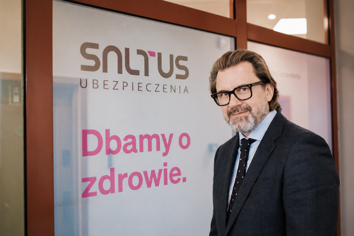 Robert Łoś, Prezes Zarządu SALTUS Ubezpieczenia, stojący w okularach i garniturze na tle szklanych drzwi z logo SALTUS Ubezpieczenia i hasłem Dbamy o zdrowie