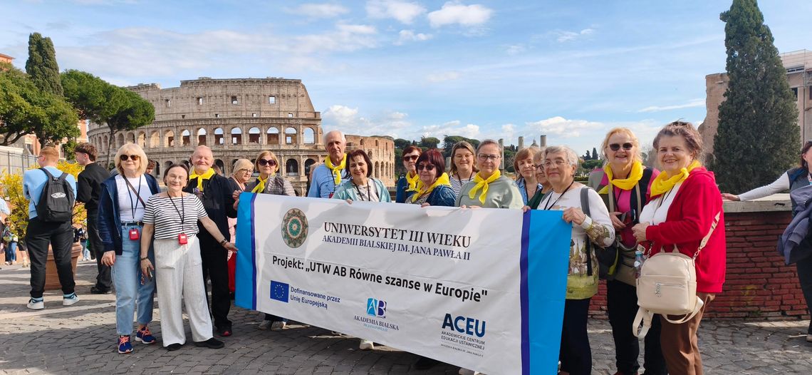 Grupa seniorów stoi na rzymskim placu, trzymając duży biały baner z logo Akademii Bialskiej i napisem „UTW AB Równe szanse w Europie”. W tle widać monumentalną, antyczną architekturę Koloseum pod błękitnym niebem. Uczestnicy mają na szyjach charakterystyczne żółte chusty.