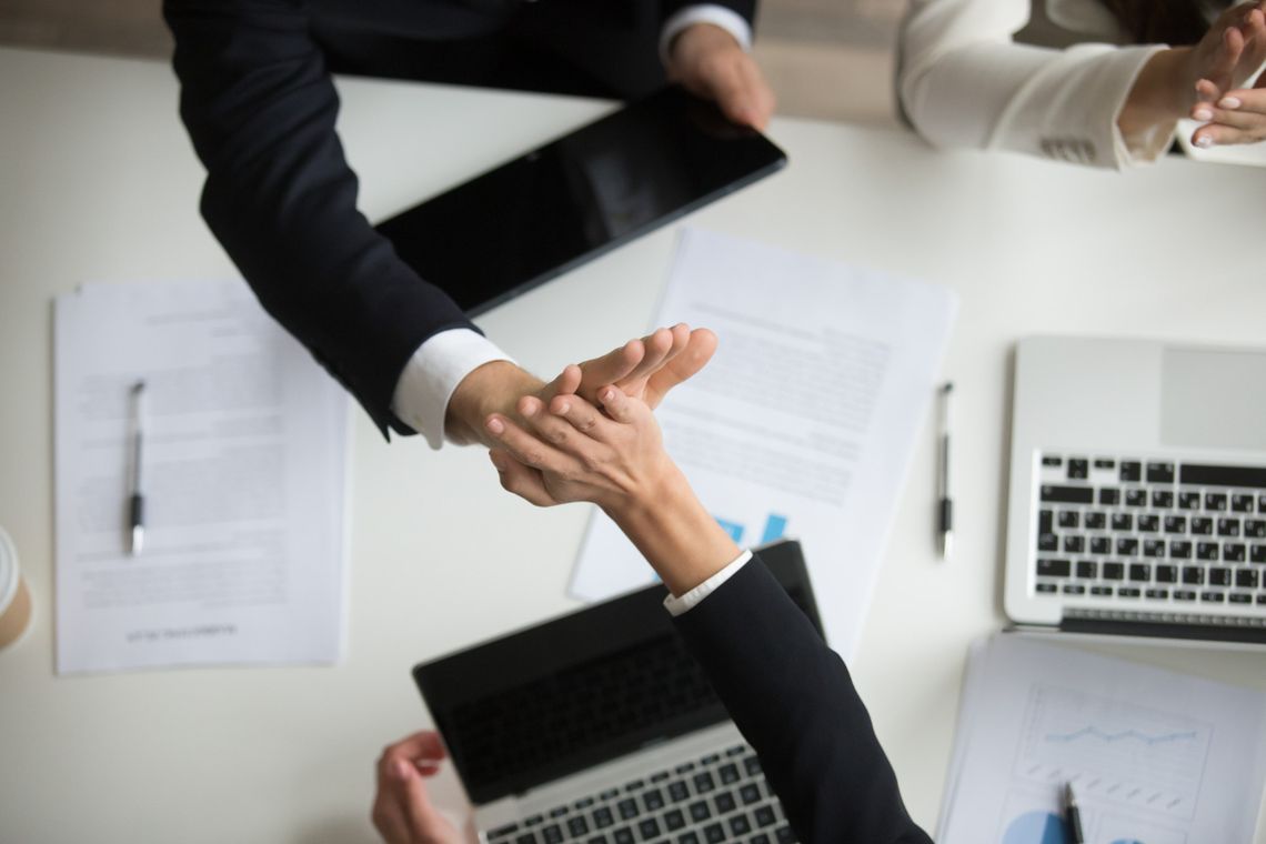 Widok z góry na biurko, przy którym dwie osoby w biznesowych garniturach przybijają „piątkę” nad otwartymi laptopami i dokumentami. Zdjęcie symbolizuje udaną rekrutację, porozumienie oraz pozytywną energię w pracy, promując cykl „Pracowity wtorek” w Słowie Podlasia.