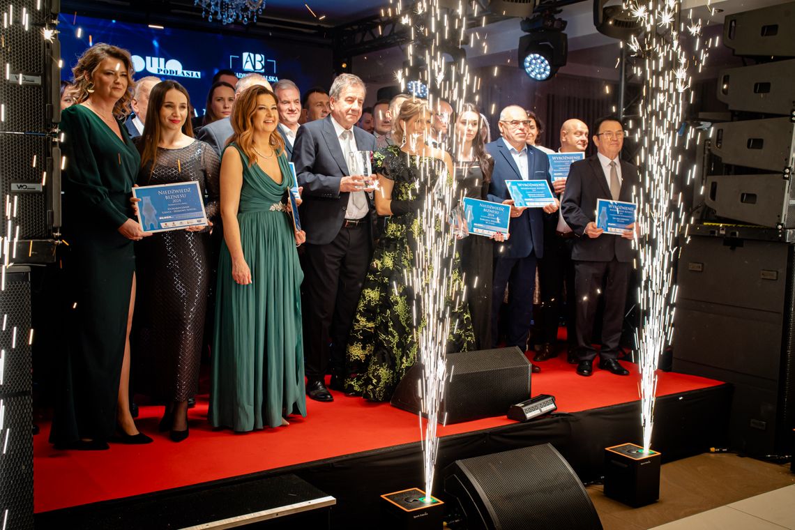 Twórzmy razem historię sukcesu naszego regionu! Wystartował Plebiscyt Gospodarczy „Niedźwiedzie Biznesu 2026” Grupa elegancko ubranych kobiet i mężczyzn stoi na scenie z czerwonym dywanem, trzymając w rękach dyplomy i statuetki. W tle widać logo Słowa Podlasia, a na pierwszym planie z podłogi strzelają dwa efektowne, iskrowe fontanny (zimne ognie).