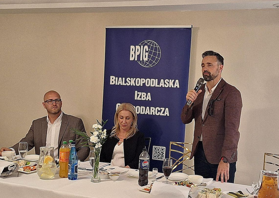 Walne Zgromadzenie BPIG: Podsumowanie pracowitego roku i 35-lecia bialskiej izby Trzy osoby przy stole prezydialnym podczas oficjalnego spotkania. Po prawej stronie stoi mężczyzna w brązowej marynarce z mikrofonem w ręku i przemawia. Na środku siedzi blondynka w czarnym żakiecie, a po lewej stronie siedzi mężczyzna w okularach i szarej marynarce.
