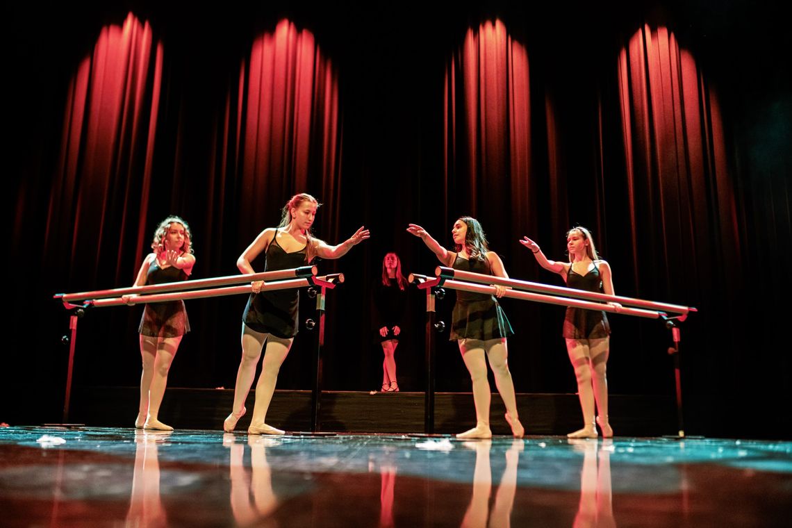 Z Białej Podlaskiej na światowe sceny. Rekrutacja do Niepublicznej Szkoły Sztuki Tańca Caro Dance w Siedlcach Cztety młode tancerki w czarnych kostiumach i puentach ćwiczą przy drążkach baletowych na tle czerwonej scenicznej kurtyny.