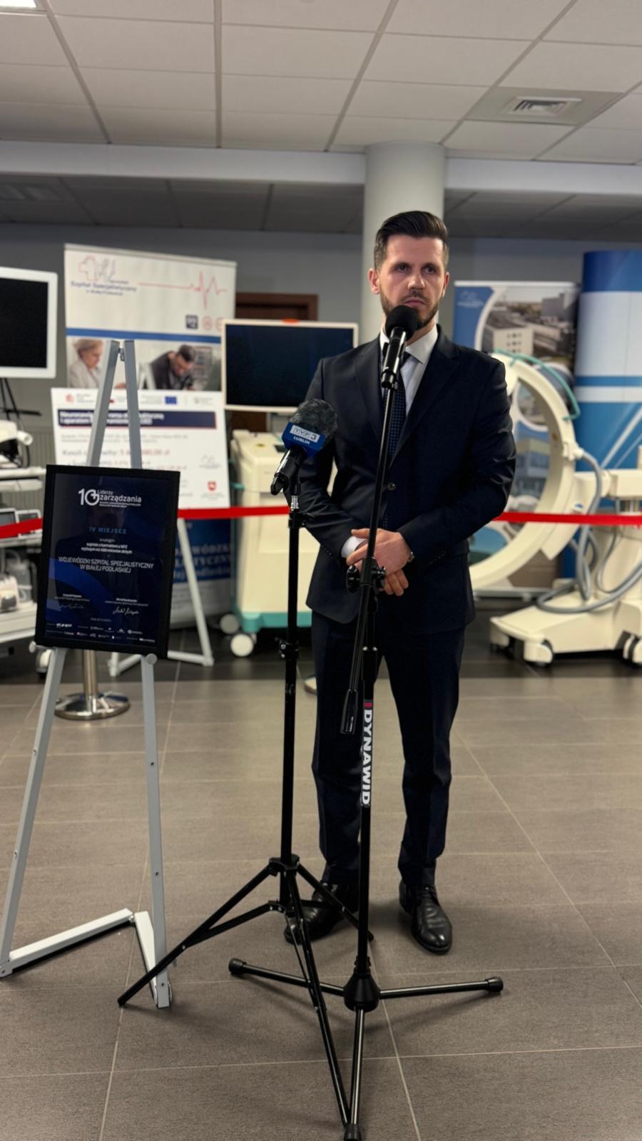 Dyrektor Wojewódzkiego Szpitala Specjalistycznego w Białej Podlaskiej, Artur Kozioł, stoi przy mikrofonie podczas oficjalnego wystąpienia. Obok na sztaludze widoczna jest oprawiona pamiątkowa tablica potwierdzająca zajęcie IV miejsca w 10. edycji rankingu Liderzy Zarządzania.