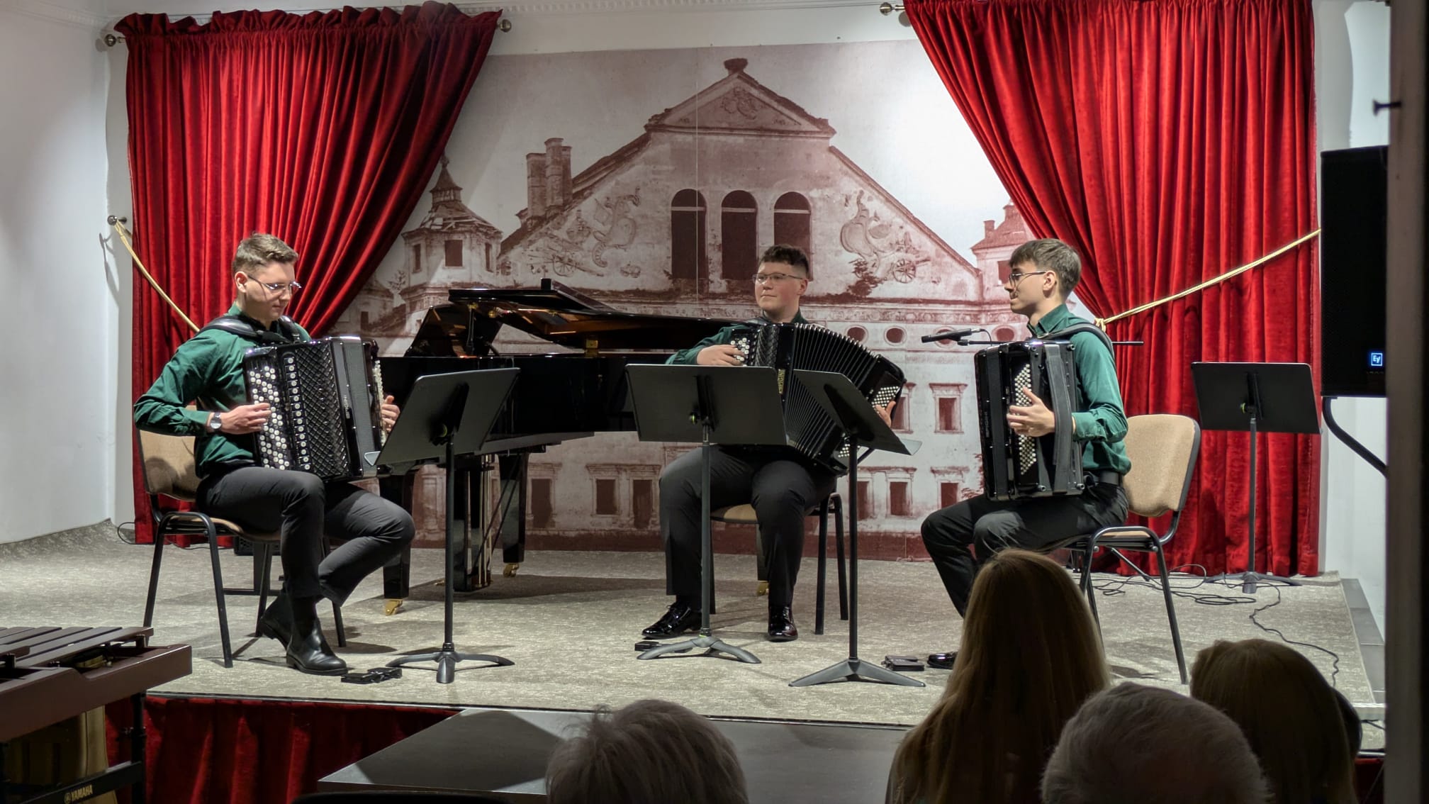 Trio akordeonowe na scenie. Trzech młodych muzyków w zielonych koszulach siedzi i gra na akordeonach. W tle czarny fortepian, czerwone kotary oraz grafika z zabytkową architekturą. Na pierwszym planie widoczne głowy publiczności siedzącej w Sali Koncertowej.