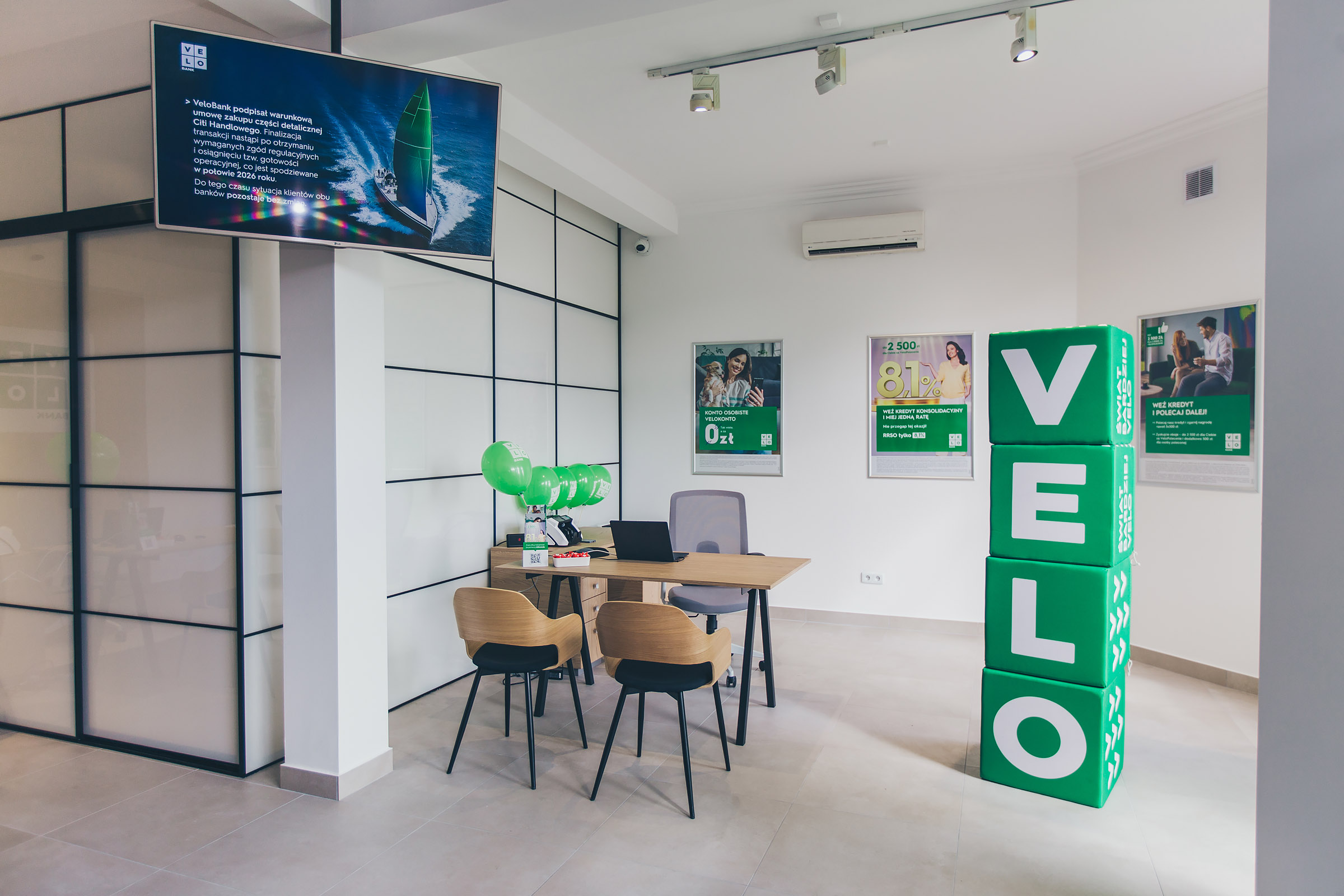 Widok z zewnątrz na nową placówkę VeloBanku w Białej Podlaskiej. Przeszklone wejście do lokalu przy ul. Gabriela Narutowicza 40 z wyraźnym, podświetlonym logo VeloBank nad drzwiami. Witryny oklejone są grafikami promocyjnymi banku w kolorach białym i zielonym.