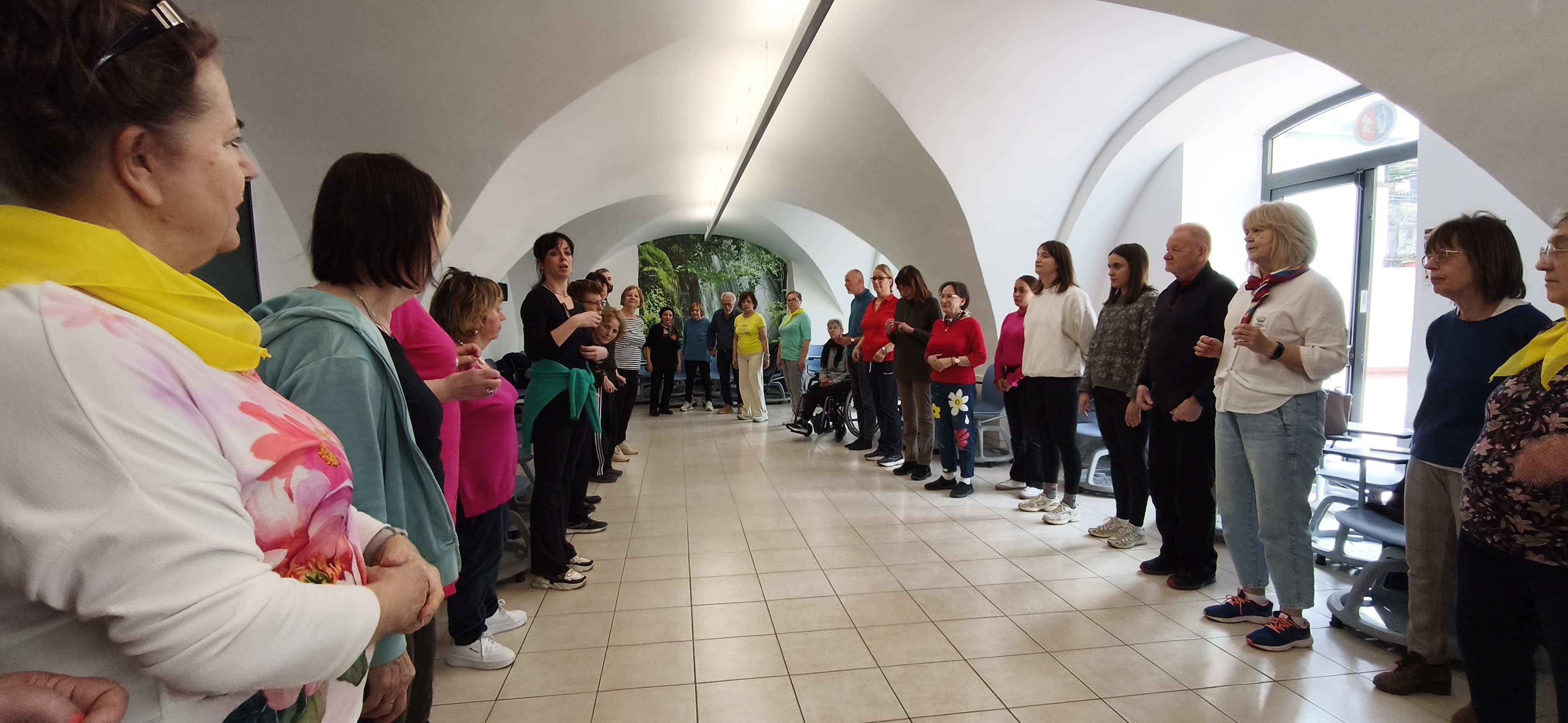 Grupa seniorów i edukatorów stoi w dwóch rzędach naprzeciw siebie w jasnej sali o sklepieniu kolebkowym. Uczestnicy biorą udział w zajęciach ruchowych, patrząc na instruktorkę. Panuje atmosfera skupienia i współpracy. Niektórzy mają na szyjach żółte chusty identyfikujące grupę z Polski.