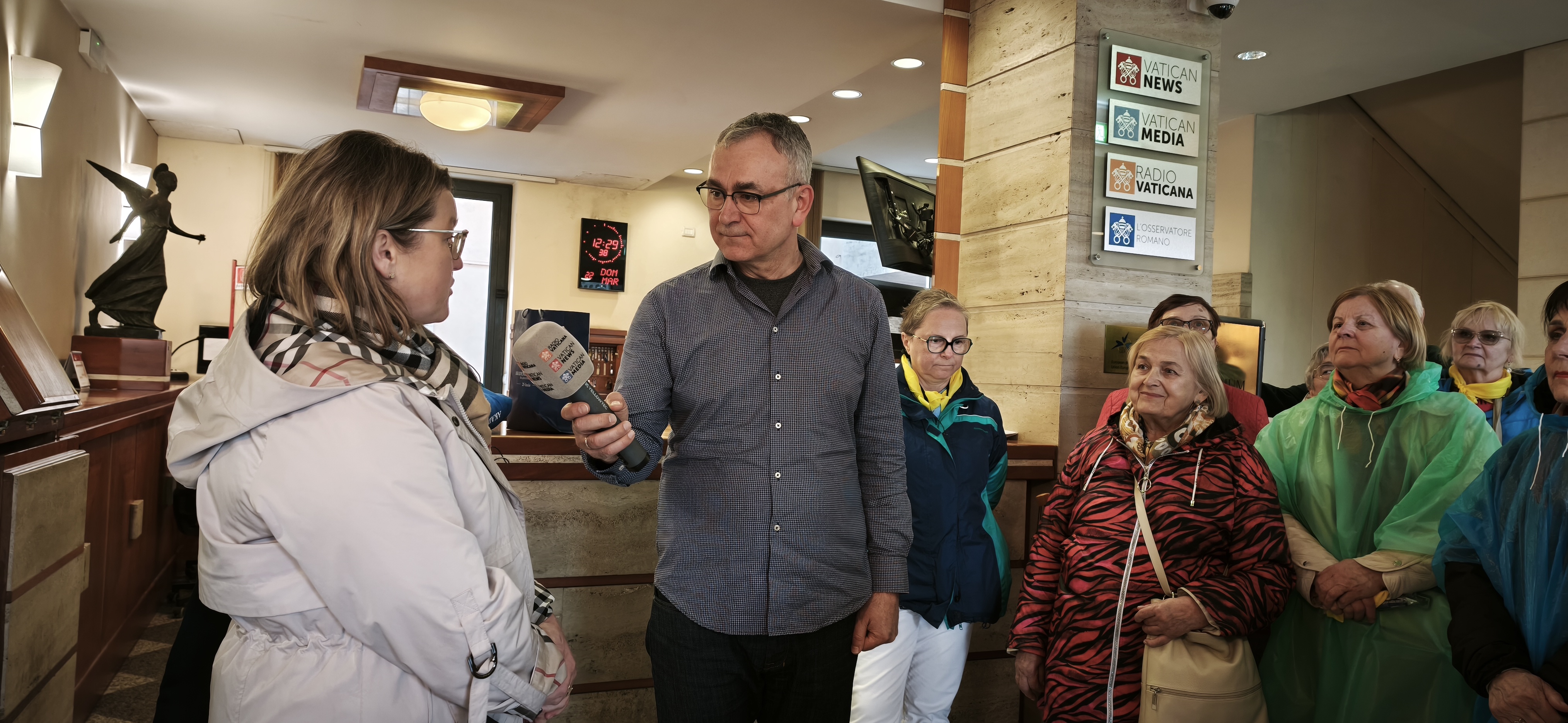 Mężczyzna w okularach trzyma mikrofon z logo Vatican News, przeprowadzając wywiad z kobietą w jasnym płaszczu. W tle widać grupę uśmiechniętych seniorek w kolorowych kurtkach i pelerynach przeciwdeszczowych. Na ścianie po prawej widnieją tablice informacyjne "Radio Vaticana".