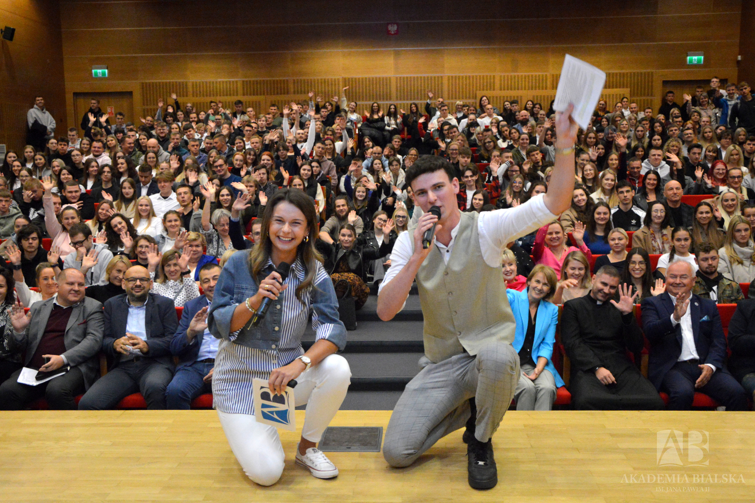 Dwoje uśmiechniętych studentów (kobieta i mężczyzna w kamizelce) klęczy na scenie auli, trzymając mikrofony i logotyp Akademii Bialskiej. Za nimi, na widowni, setki osób machają rękami w stronę aparatu. W tle widać logo uczelni.