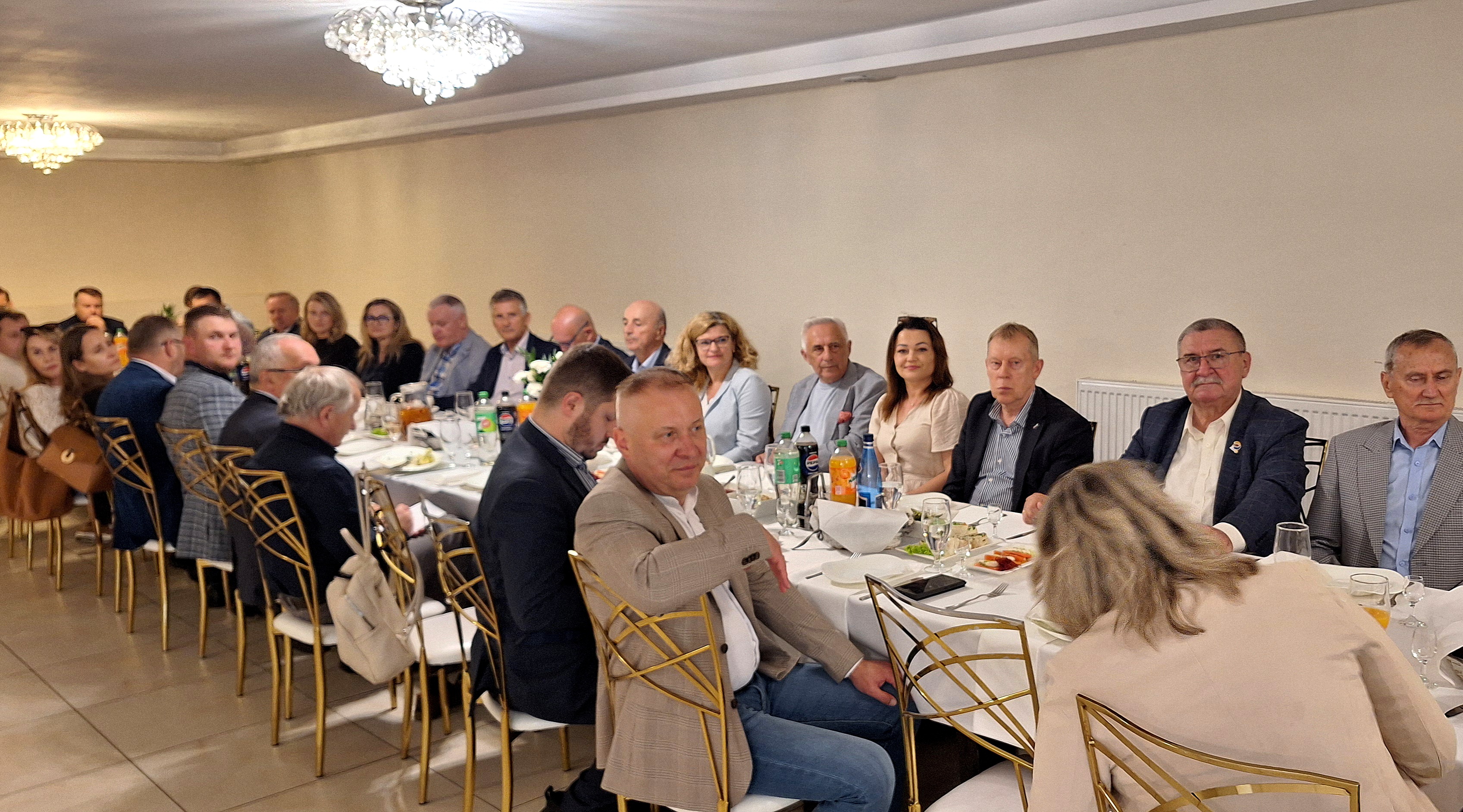 Szerokie ujęcie eleganckiej sali bankietowej, w której uczestnicy oficjalnego spotkania siedzą przy długim, zastawionym stole ułożonym w kształcie litery U. Widoczna jest liczna grupa kobiet i mężczyzn w strojach biznesowych, siedzących na ozdobnych, złotych krzesłach z białymi obiciami. Na stołach pokrytych białymi obrusami znajdują się talerze z potrawami, napoje oraz szklanki. Wnętrze jest jasne, oświetlone przez ozdobne żyrandole sufitowe.
