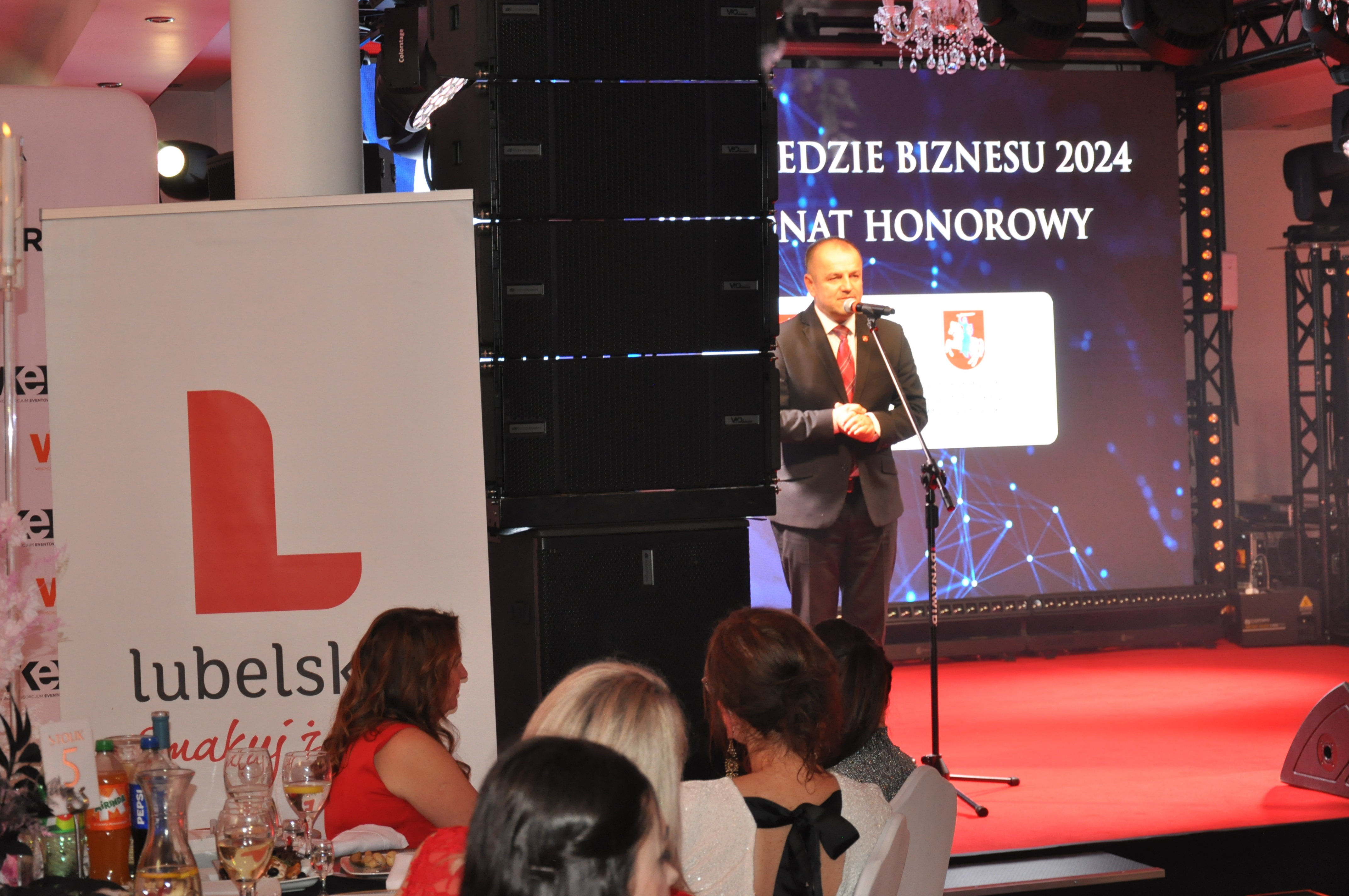 Dariusz Litwiniuk, Kierownik Filii Urzędu Marszałkowskiego w Białej Podlaskiej, przemawia na scenie podczas Gali Niedźwiedzie Biznesu. W tle widoczny telebim z herbem województwa, a na pierwszym planie goście przy stolikach oraz materiały promocyjne.